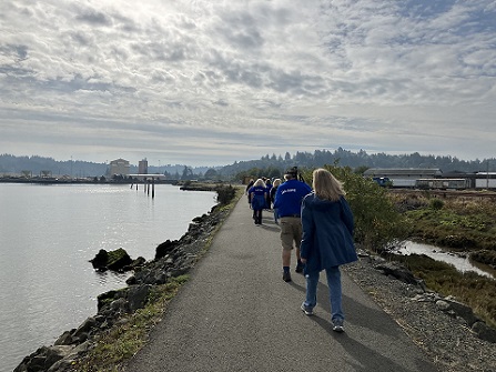 Pic of Sole Mates walking Coos Bay Boardwalk trail. Pic taken from behind walkers showing their backs w/SE side of town and cloads in background