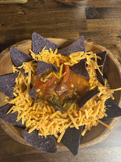 Pic of Josh's Vegetable Tortilla Soup in bowl on wood table w/bluw corn chips and grated cheddar chees, etc.