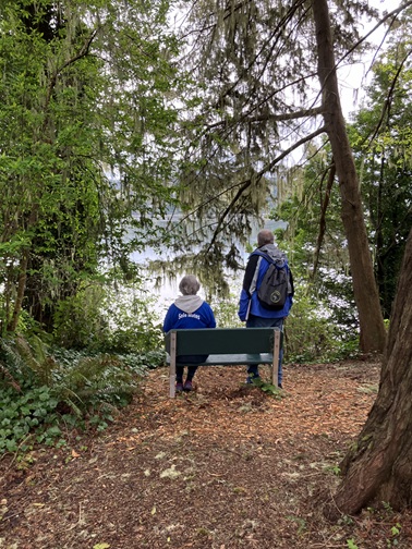 Two Sole Mates with backs to camera at bench on Ferry Road Park Trail,. Man standing with backpack on, woman sitting at bench's shirt reads "Sole Mates" on back. Two are facing bay surrounded by trees.