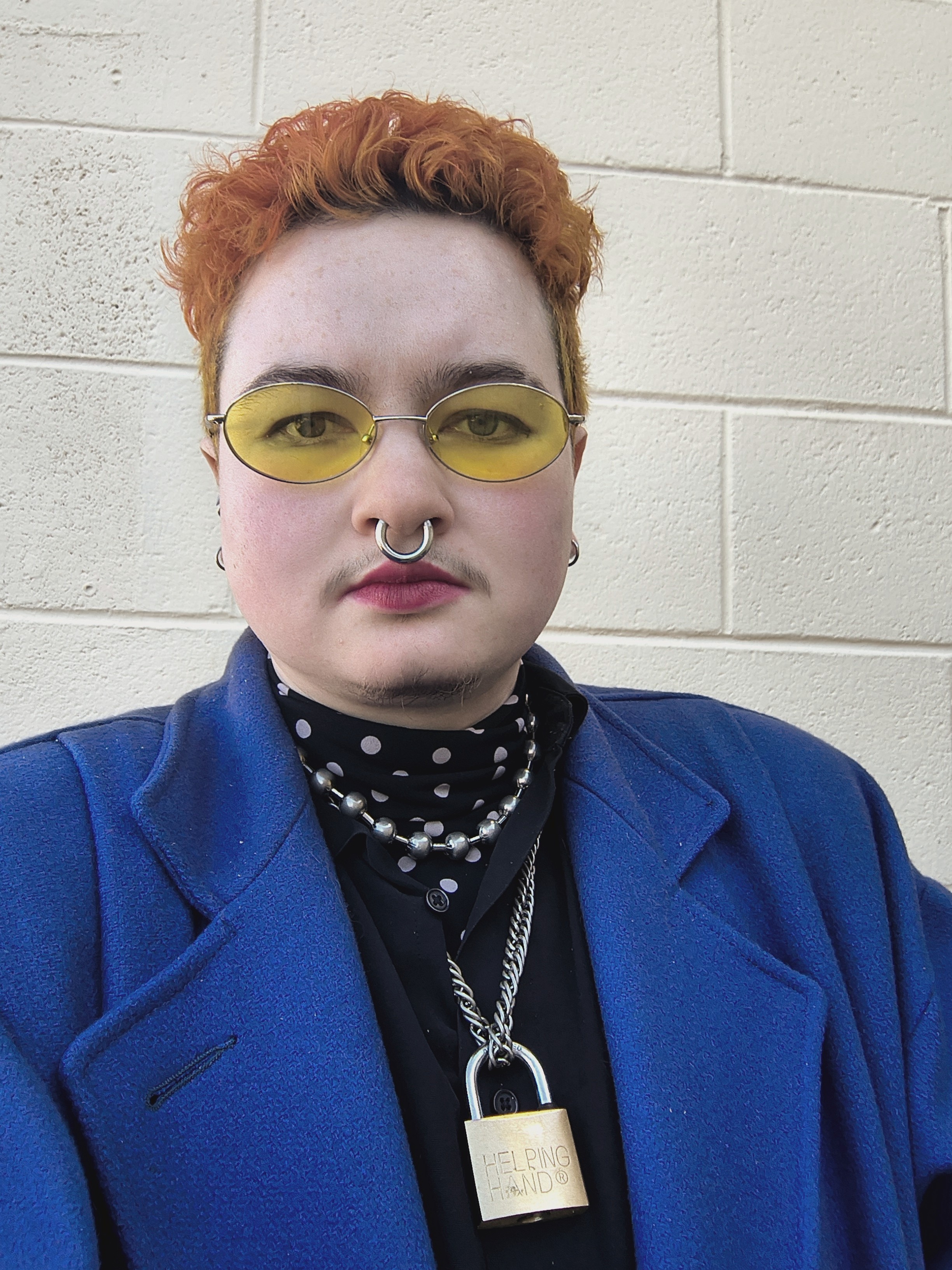 pic of Marlow with short orange hair, yellow tinted shades, blue felt coat, white brick wall in background