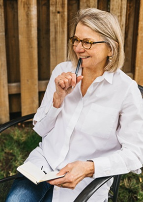Waterston sitting in chair in yard with wood fence behind her. She's wearing a white button up and is holding a pen to her mouth, smiling, with notebook in her lap