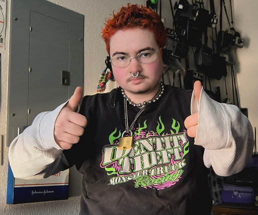 Pic of Marlow in room, with old cameras hanging on wall in background, wearing white thermal under black Identity Theft Monster Truck t-shirt, with thums up